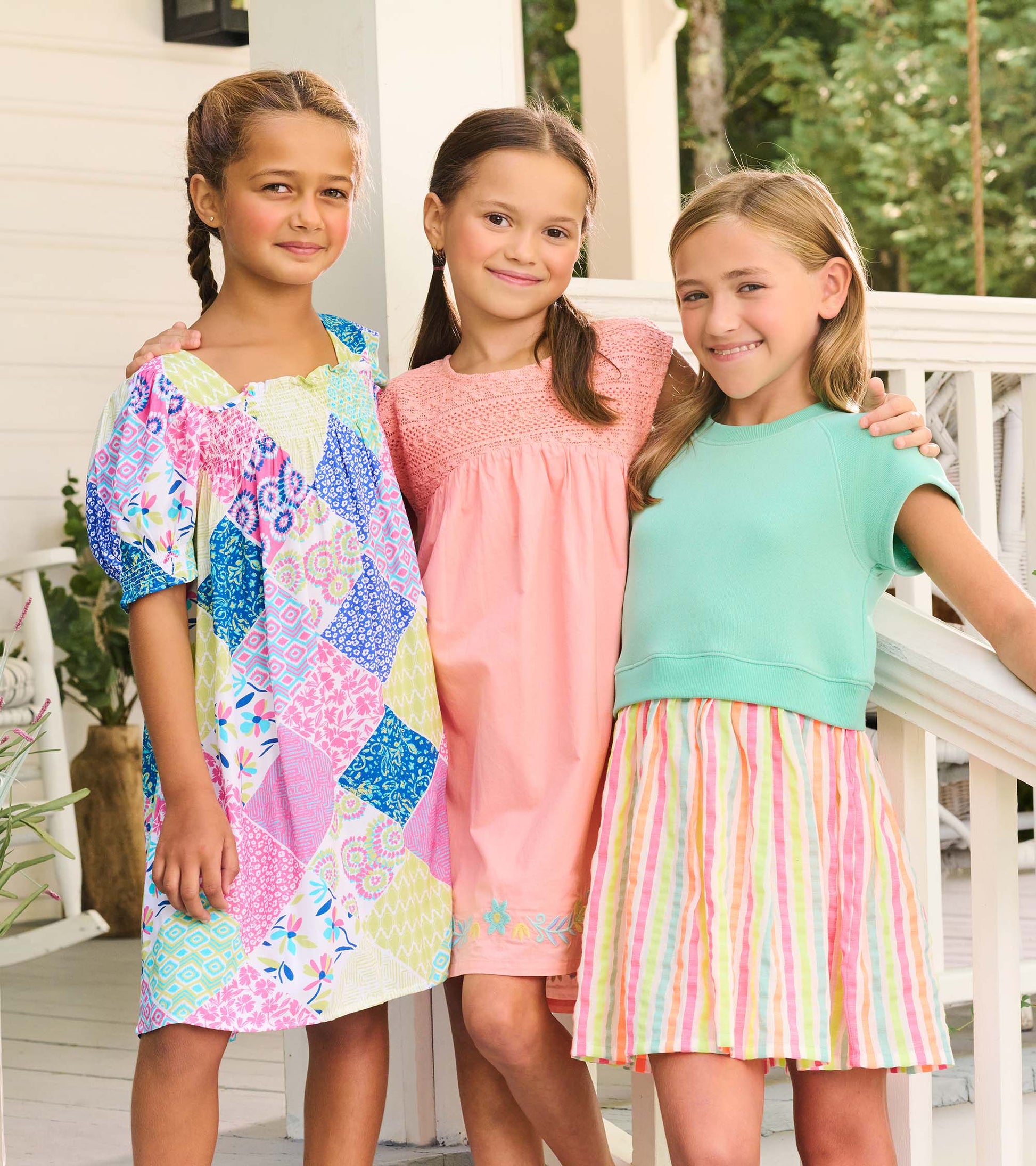 Three girls wearing colorful summer dresses, including a patchwork print dress, a pink dress, and a mint top with striped sk…