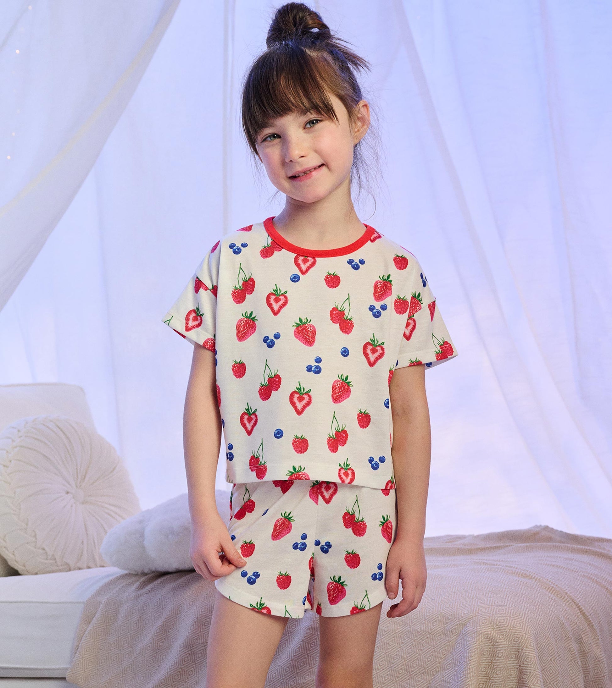 Girl wearing cream short pajamas with berry print and red neckline trim, standing front view