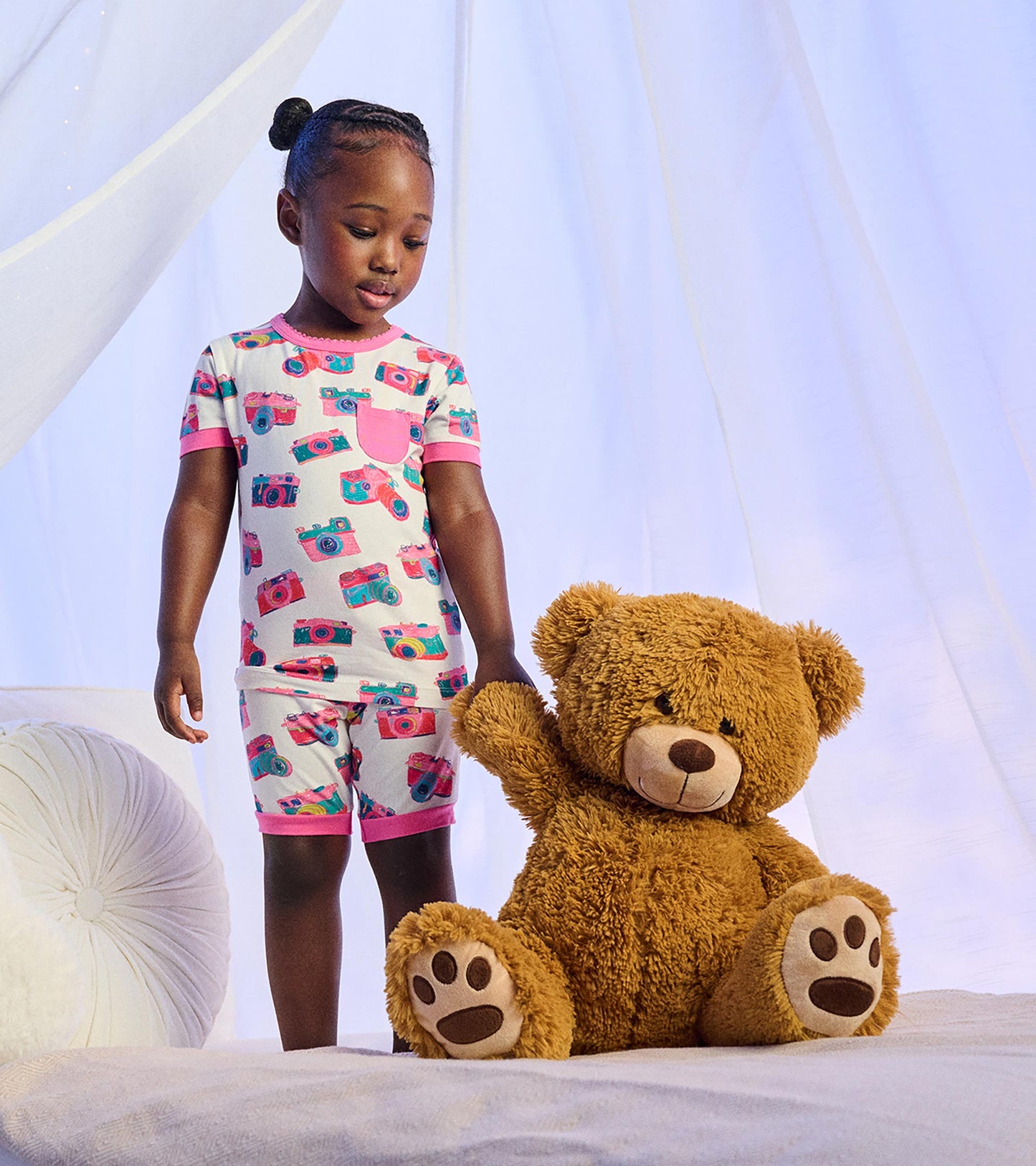 Girl wearing white short pajamas with colorful camera print and pink trim, standing beside a large teddy bear