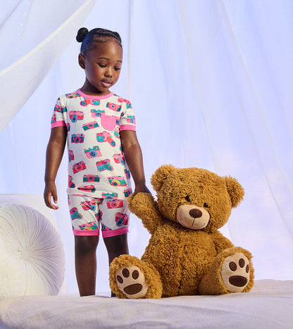 Girl wearing white short pajamas with colorful camera print and pink trim, standing beside a large teddy bear