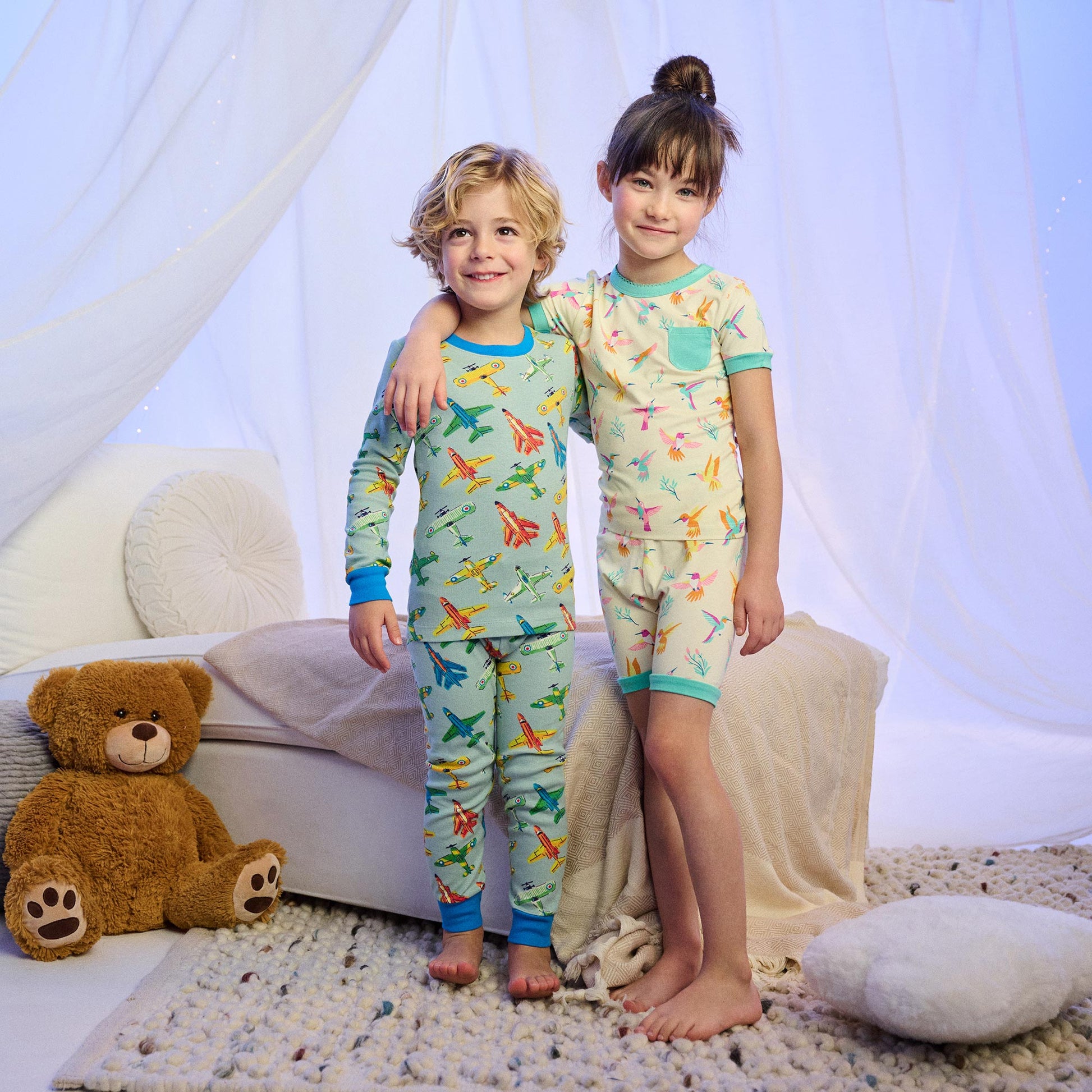 Two children wearing printed bamboo pajama sets, one with airplanes and one with hummingbirds, standing indoors.