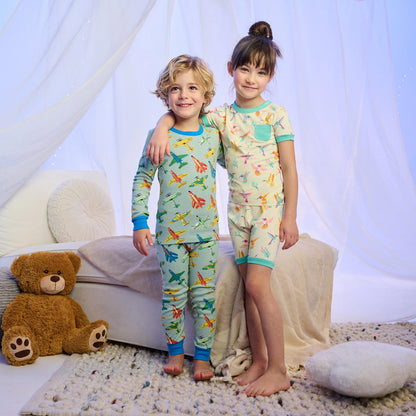 Two children wearing printed bamboo pajama sets, one with airplanes and one with hummingbirds, standing indoors.
