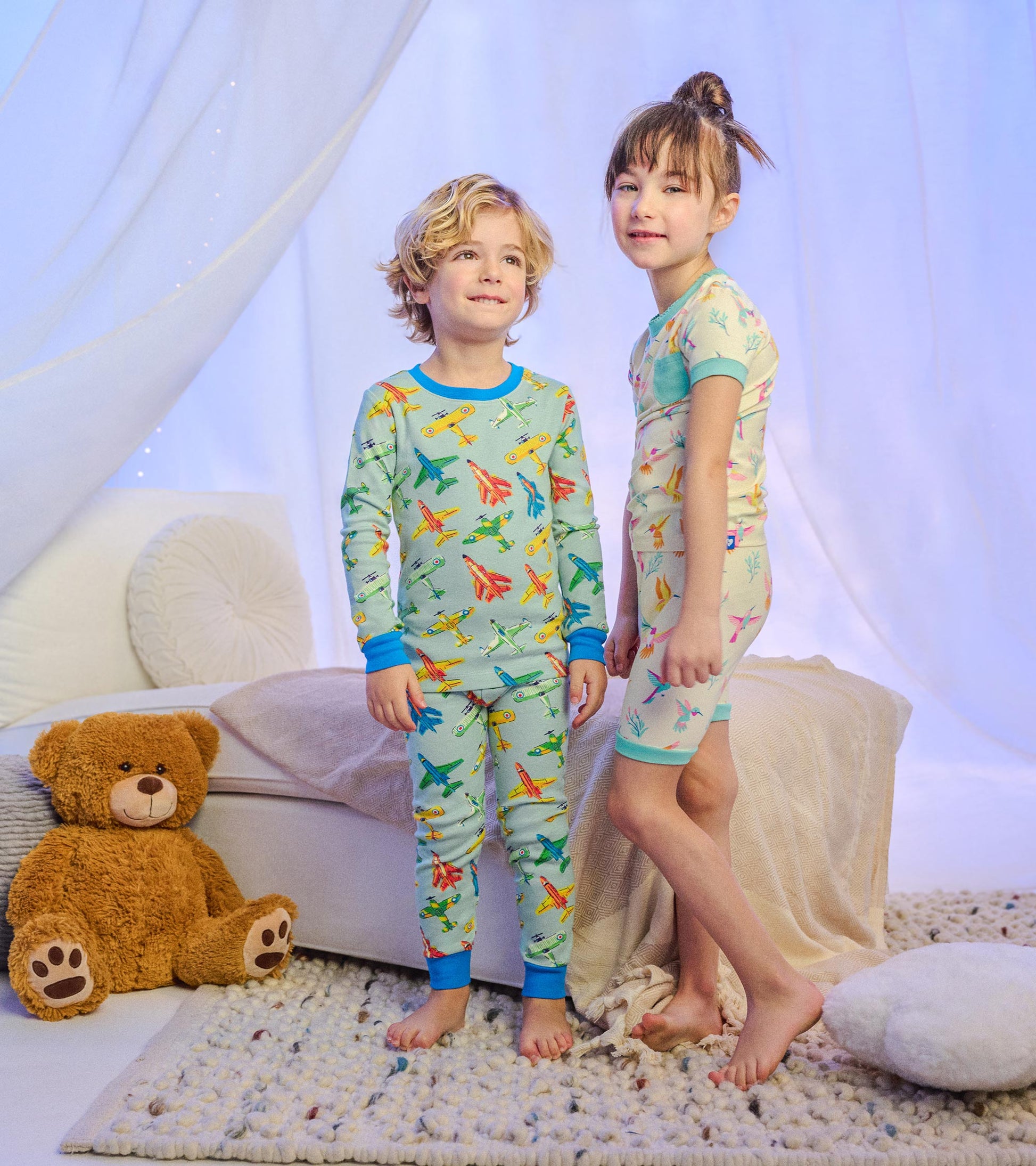 Two children wearing long-sleeve bamboo pajamas, one blue airplane print and one cream hummingbird print, standing indoors.