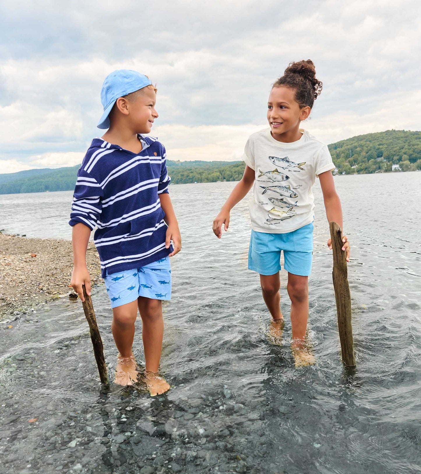 Two kids in water wearing graphic tees, one cream fish print tee and one navy striped hooded tee
