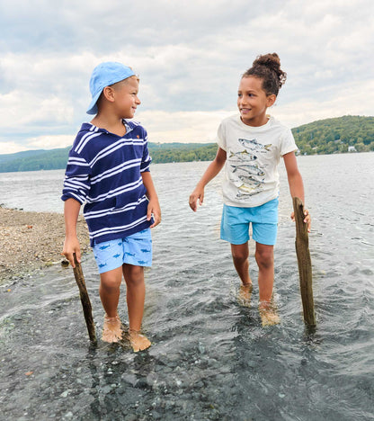 Two kids in water wearing graphic tees, one cream fish print tee and one navy striped hooded tee