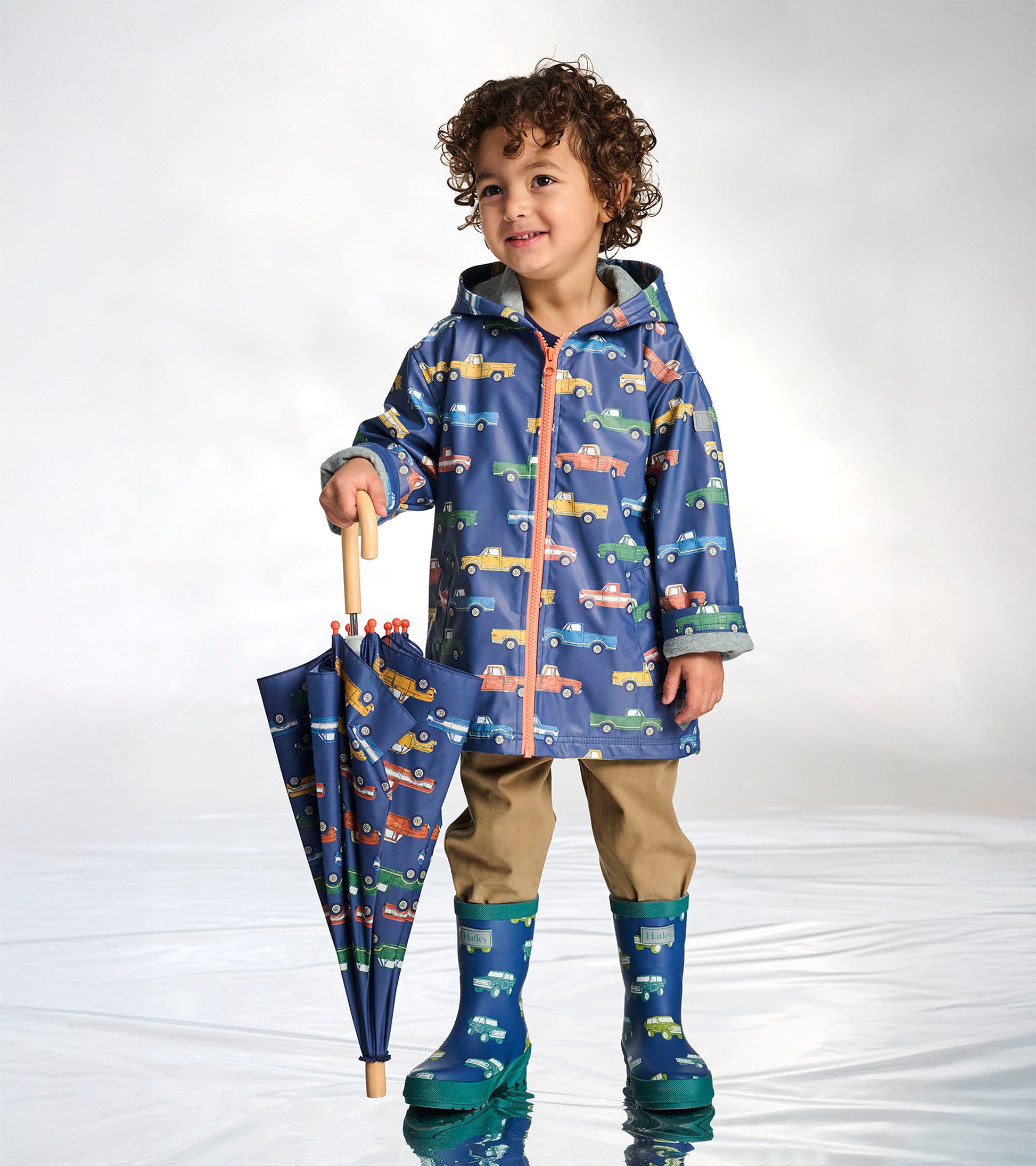Boy wearing a navy zip-up raincoat with colorful truck print and coral zipper, holding a matching umbrella