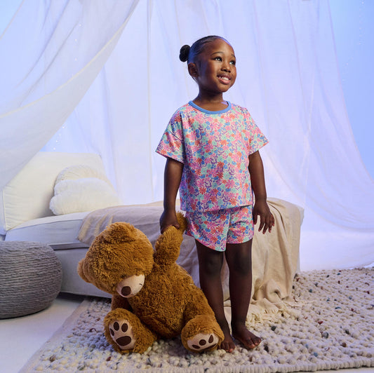 Girl wearing floral short pajama set with blue trim, standing barefoot beside a teddy bear