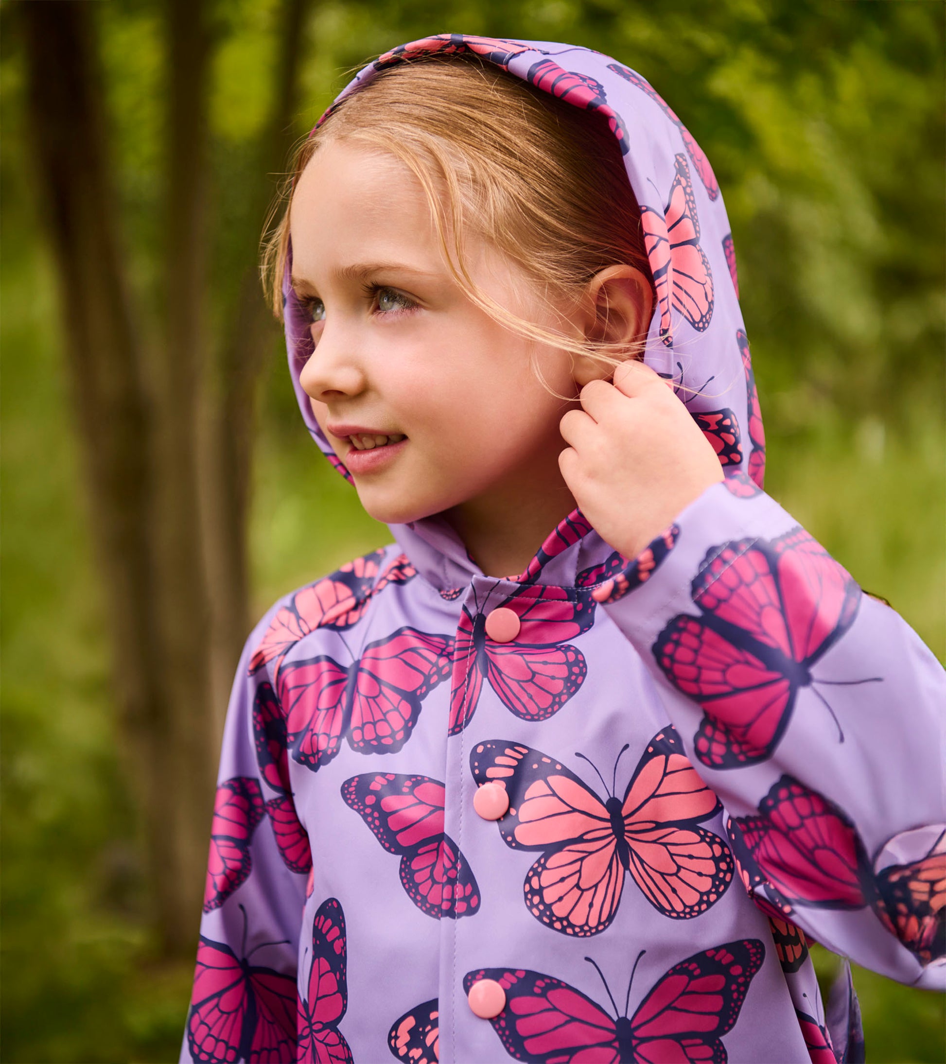 Girl wearing a lavender hooded raincoat with pink butterfly print and snap buttons, front view
