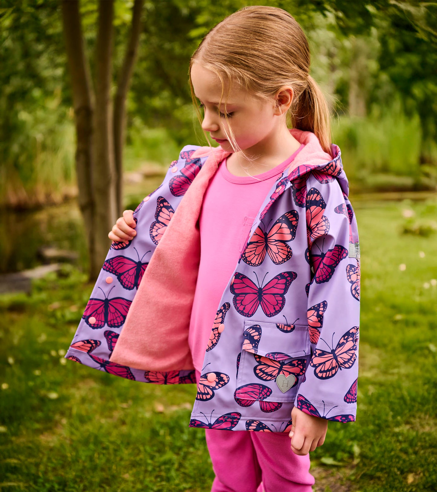 Lavender hooded girls raincoat with pink butterfly print, pink lining, and front pockets, worn open outdoors