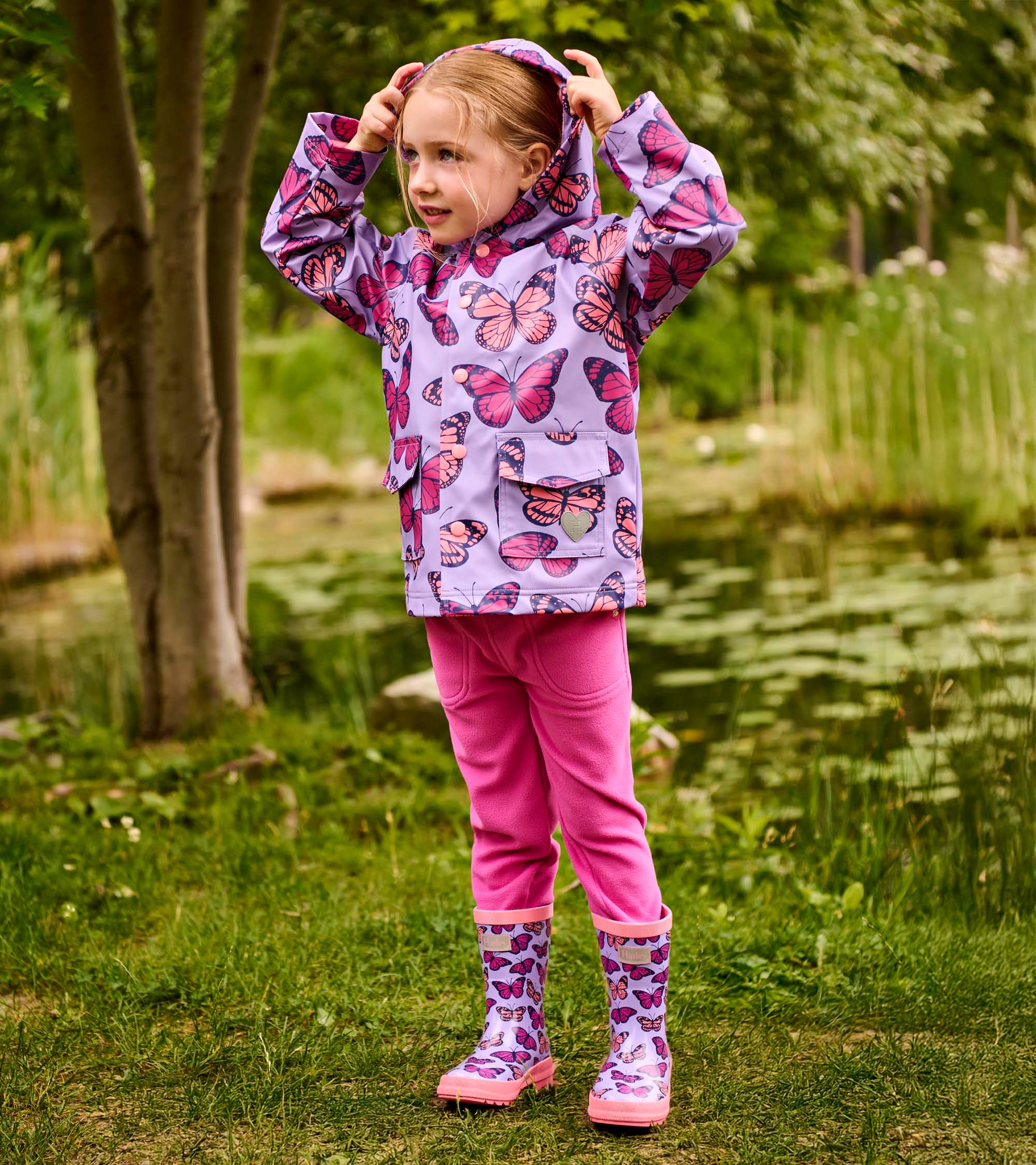 Girl wearing a lavender butterfly-print hooded raincoat with pink snaps and pockets, front view