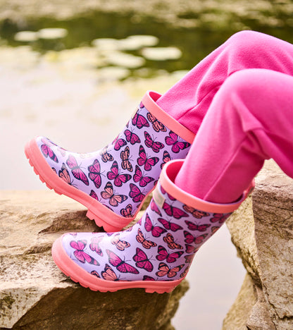 Lavender kids rain boots with pink butterfly print and pink trim, shown on feet outdoors
