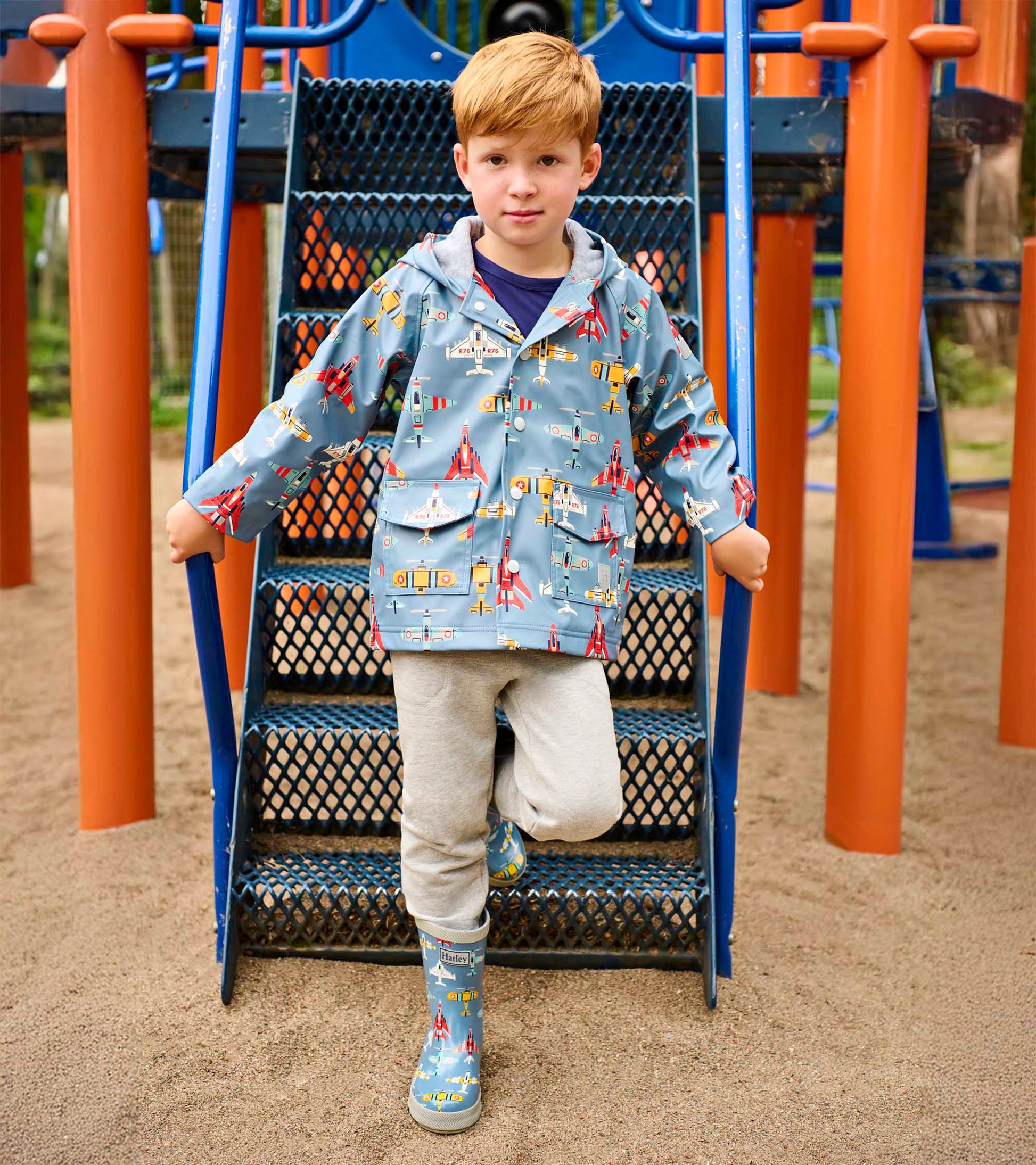 Light blue boys raincoat with airplane print, hood, front pockets, and snap closure, worn by a child outdoors