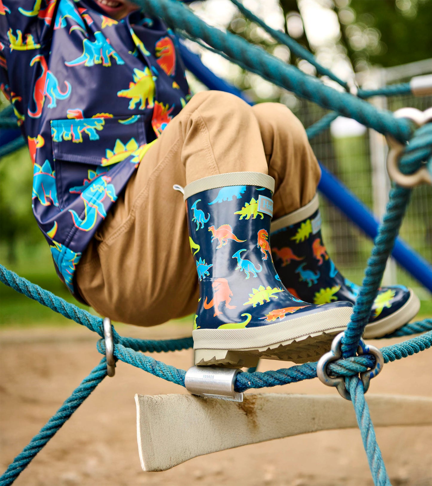 Navy kids rain boots with colorful dinosaur print and beige soles, shown on a child climbing ropes