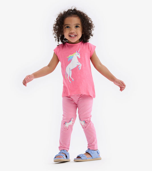 Pink toddler tee with unicorn graphic, paired with striped leggings featuring unicorns, worn with blue sandals.
