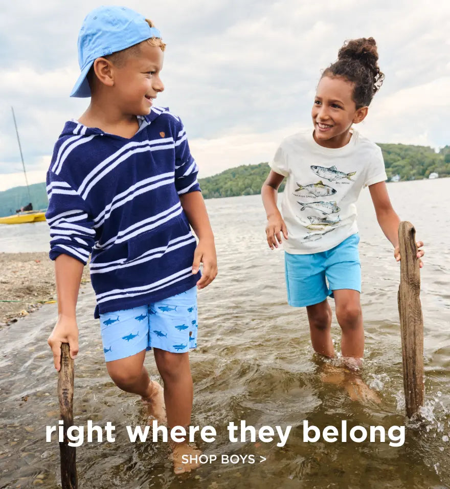 Shop boys' clothing. Showing two children wading and smiling by a lakeshore in casual summer outfits.