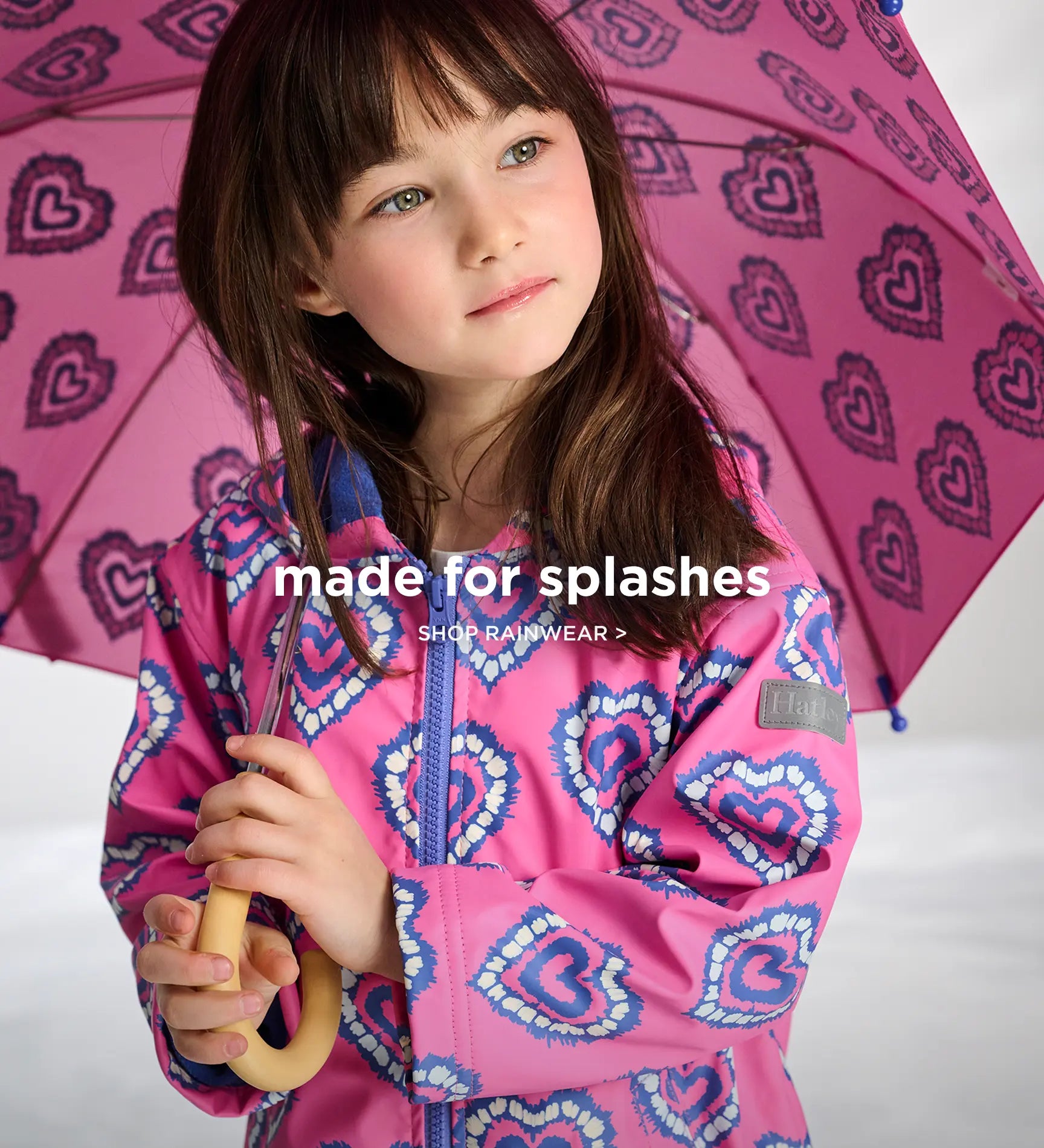 Promotes the kids' rainwear collection. Showing girl in a pink heart-print raincoat holding a matching umbrella.