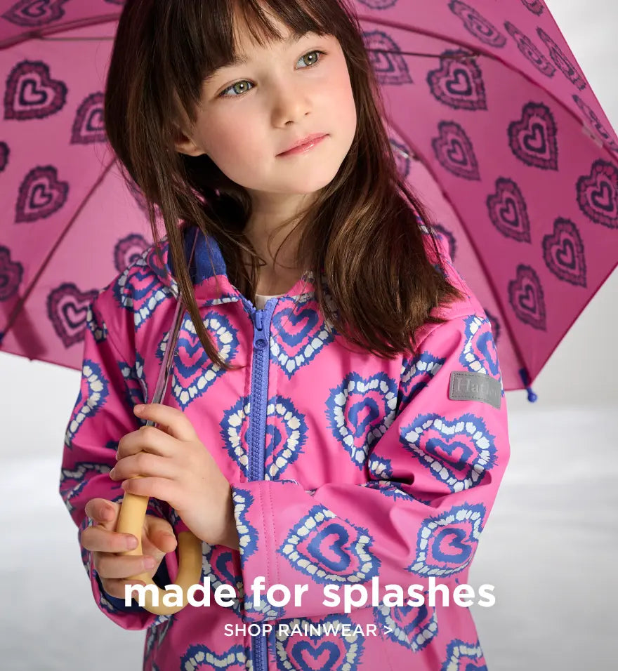Shop kids' rainwear. Showing girl in a pink heart-print raincoat holding a matching umbrella.